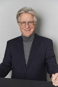 Das Bild steht in Zusammenhang mit Mediation und Konfliktlösung durch die Kanzlei Ponschab & Partner und zeigt folgendes: Ein älterer Mann mit grauen Haaren und Brille trägt ein dunkles Sakko und einen grauen Rollkragenpullover. Er lächelt und steht vor einem schlichten, hellen Hintergrund. Bei Fragen zu Konflikten und Konfliktlösung wenden Sie sich bitte an uns unter info@ponschab-partner.com