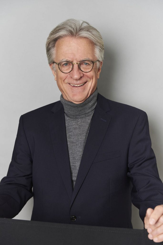 Das Bild steht in Zusammenhang mit Mediation und Konfliktlösung durch die Kanzlei Ponschab & Partner und zeigt folgendes: Ein älterer Mann mit grauen Haaren und Brille trägt ein dunkles Sakko und einen grauen Rollkragenpullover. Er lächelt und steht vor einem schlichten, hellen Hintergrund. Bei Fragen zu Konflikten und Konfliktlösung wenden Sie sich bitte an uns unter info@ponschab-partner.com